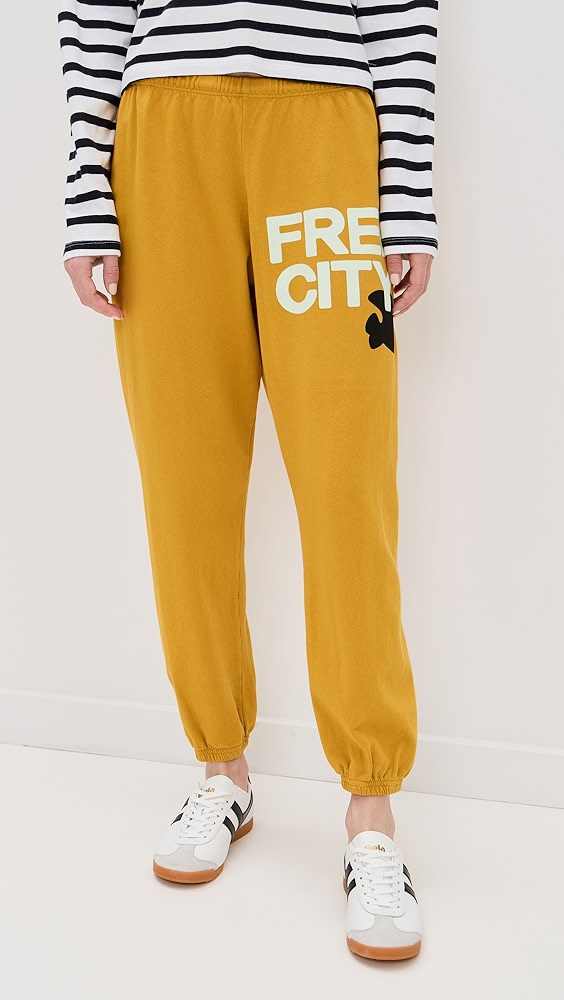 Mustard yellow sweatpants with white "FREE CITY" text and a black bird logo on the leg on a model.